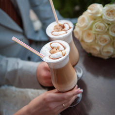 Duas pessoas a brindar com bebidas cremosas junto a um ramo de rosas