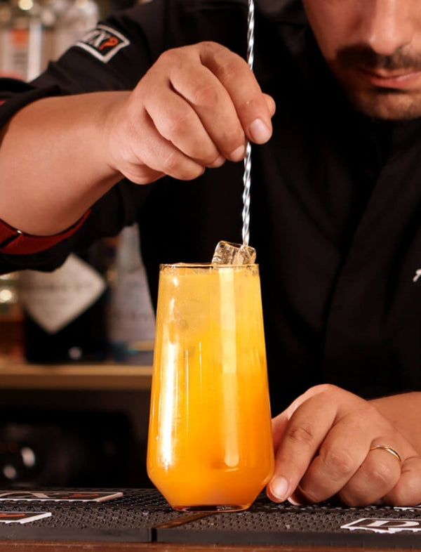 Barman a preparar um cocktail laranja num copo alto ao balcão de um bar