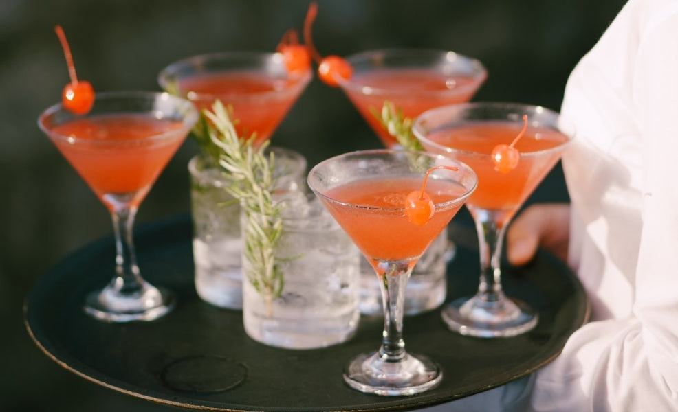 Cinco cocktails vermelhos com cereja servidos num tabuleiro em evento ao ar livre